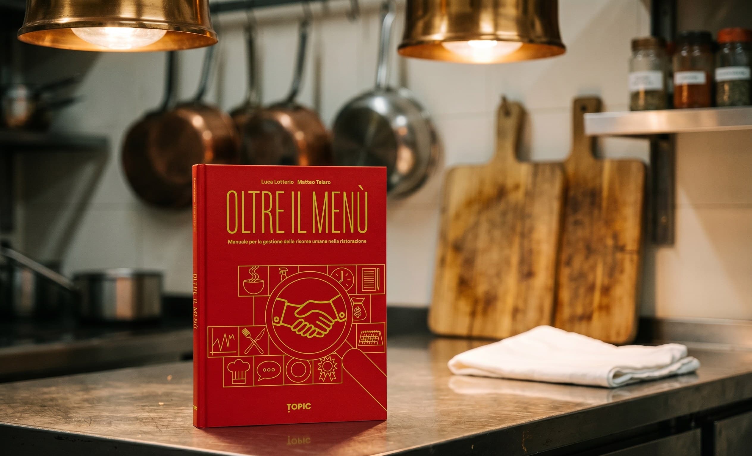 Il libro Oltre il Menù di Luca Lotterio e Matteo Telaro appoggiato su un piano di lavoro di una cucina professionale, con padelle di rame appese e taglieri sullo sfondo